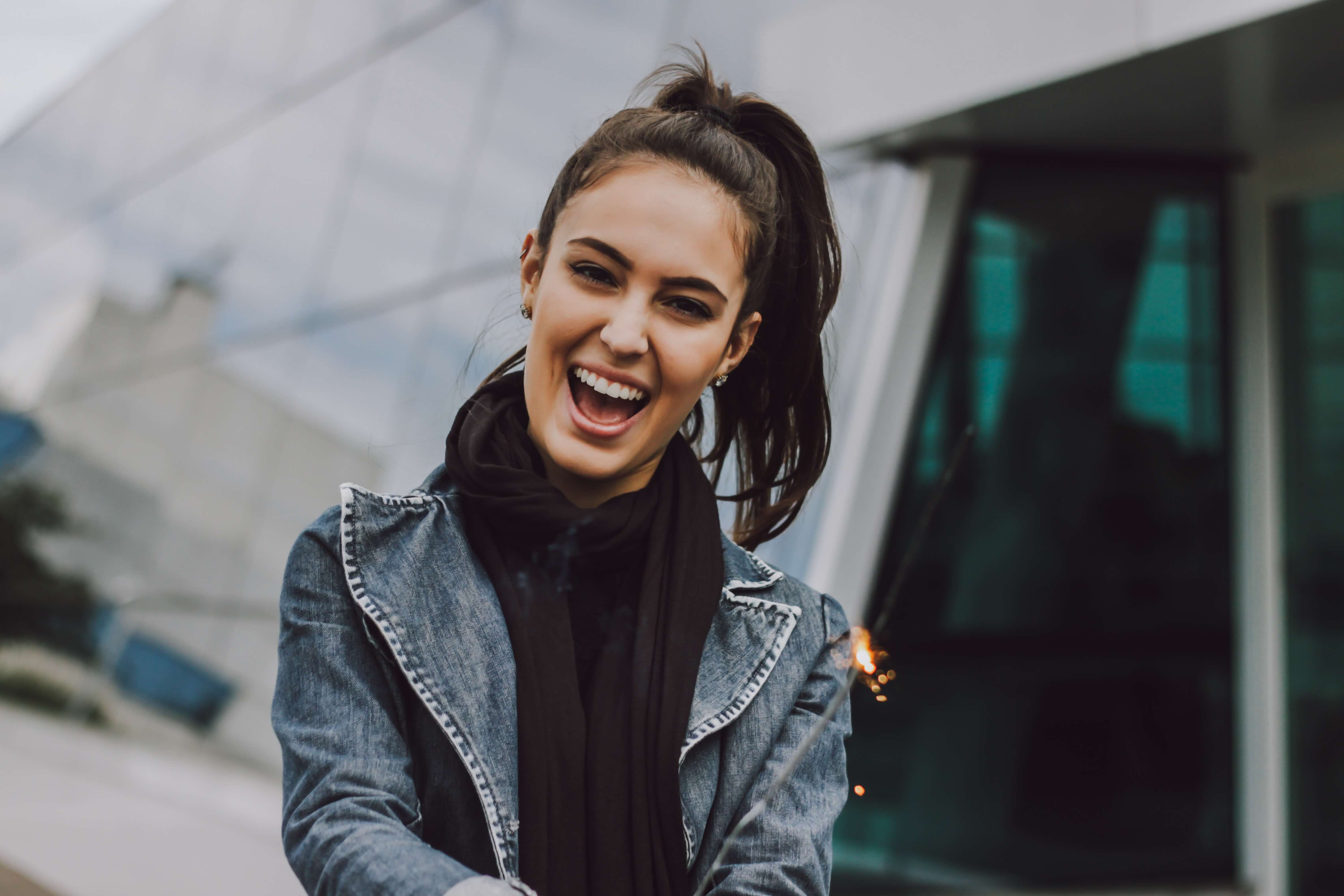A smiling woman wears a blue jean jacket in front of a glass building.