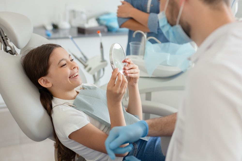 a young dental patient smiling in a mirror and laughing with her family dentist