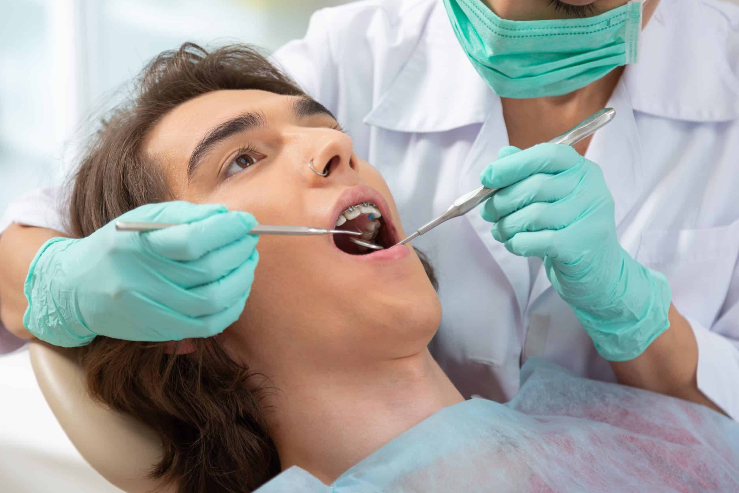 A white woman with a nose ring lays on a dentists chair with a dental bib while a dentist in a white coat with blue mask and gloves uses a mirror and scaler in her mouth.