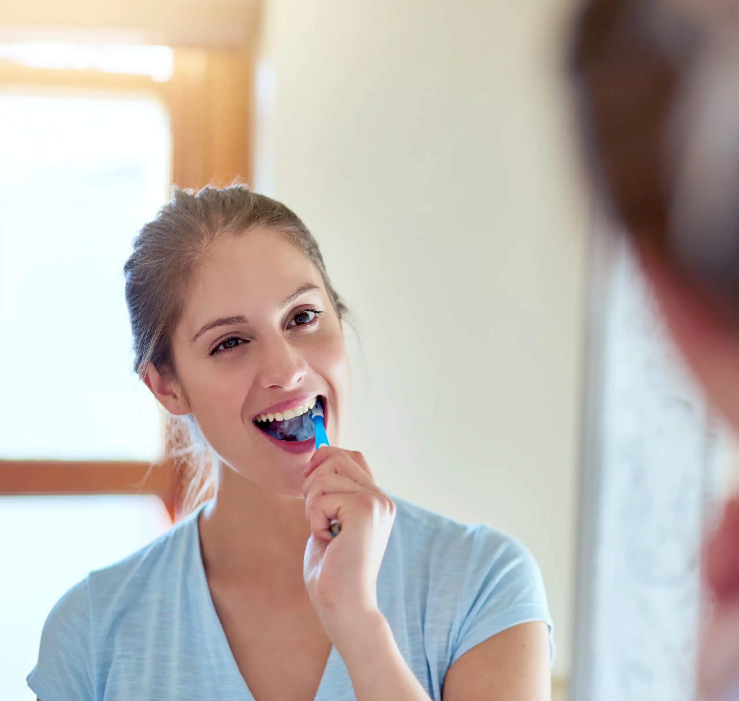 A smiling woman brushes her teeth in a mirror.