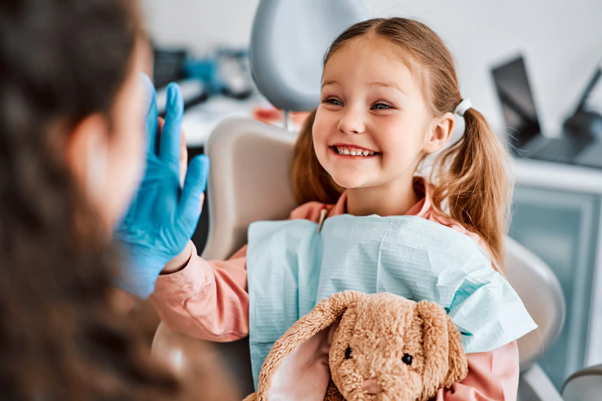 A young girl sits in a dental chair with a dental bib, holding a stuffed rabbit and high-fiving the dentist.